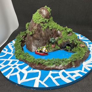 Peut inclure: Diorama miniature d'une île volcanique avec un lac et un petit bateau rouge. L'île est marron avec un feuillage vert et une grotte. Le lac est bleu et la base est un cercle bleu avec des accents blancs.