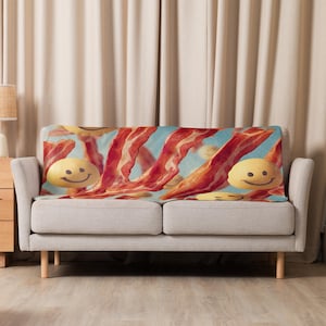 May include: A throw blanket featuring a whimsical design of bacon strips and smiling sunny-side-up eggs. The blanket is draped over a light gray sofa, with a light blue sky background. The blanket adds a playful touch to home decor.