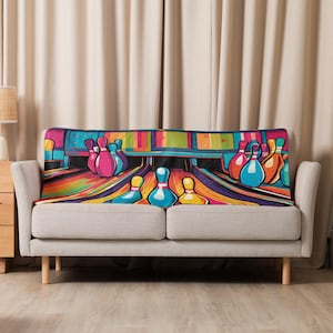 May include: A colorful bowling-themed throw blanket draped over a gray sofa. The blanket features a vibrant illustration of bowling pins and a lane in shades of pink, blue, yellow, and orange. The background includes a black area and a colorful top.