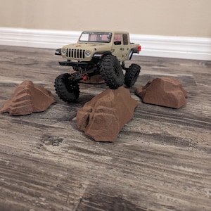 May include: A tan and black toy jeep driving over brown rock obstacles on a wood floor.