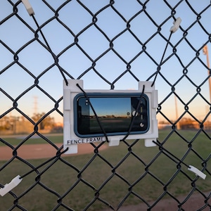 Könnte beinhalten: Ein weißer "FENCE FRAME" hält ein Smartphone an einem Maschendrahtzaun fest. Das Telefon zeigt eine Landschaft. Der Zaun ist schwarz, und der Hintergrund zeigt einen Baseballplatz unter blauem Himmel.