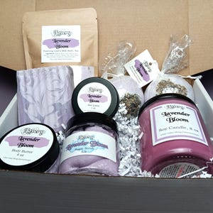 May include: A gift box filled with lavender-scented products. The box contains a soy candle, body butter, sugar scrub, aloe lotion, bath bombs, and a bag of foaming goat's milk bath. The products are in jars and containers with the "Lavender Bloom" label.