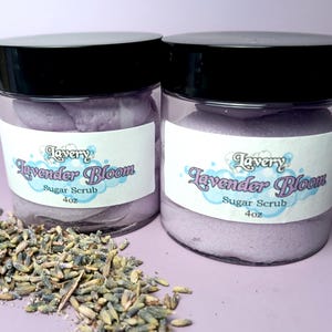 May include: Two glass jars filled with lavender-colored sugar scrub. The jars have black lids and white labels that read "Lavery Lavender Bloom Sugar Scrub 4 oz."