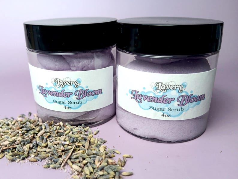 May include: Two glass jars filled with lavender-colored sugar scrub. The jars have black lids and white labels that read "Lavery Lavender Bloom Sugar Scrub 4 oz."