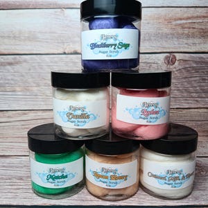 May include: Six glass jars of sugar scrub with black lids. The jars are filled with different colored scrubs, including purple, white, pink, green, orange, and white. The labels on the jars list the flavors of the scrubs: Blackberry Sage, Vanilla, Lychee, Matcha, Lemon Honey, and Oatmeal Milk & Honey. Each jar is labeled "Lavery Sugar Scrub 4 oz."