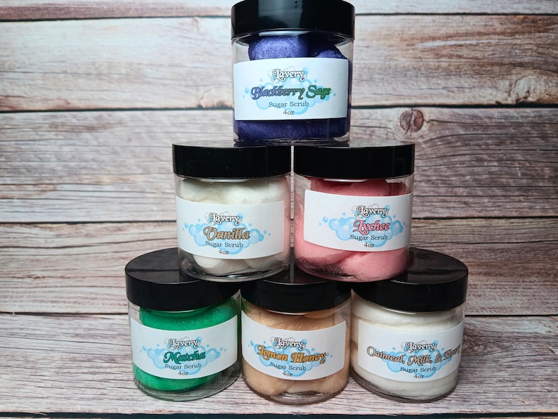 May include: Six glass jars of sugar scrub with black lids. The jars are filled with different colored scrubs, including purple, white, pink, green, orange, and white. The labels on the jars list the flavors of the scrubs: Blackberry Sage, Vanilla, Lychee, Matcha, Lemon Honey, and Oatmeal Milk & Honey. Each jar is labeled "Lavery Sugar Scrub 4 oz."