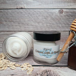May include: Two glass jars of oatmeal, milk, and honey sugar scrub. The jars are on a wooden surface with a scattering of oatmeal. The label on the jar reads "Lavery Oatmeal, Milk, & Honey Sugar Scrub 4 oz."