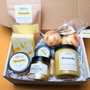 May include: A gift box filled with honeysuckle-scented bath and body products. Includes a soy candle, body butter, sugar scrub, bath bombs, soap, and foaming goat's milk bath. The products are yellow and white, with a brown paper bag and a white box.