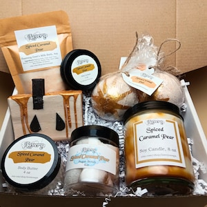 May include: A gift box filled with Spiced Caramel Pear-scented bath and body products. Includes a candle, bath bombs, body butter, sugar scrub, and a bar of soap shaped like a pumpkin. The products are in a cardboard box.