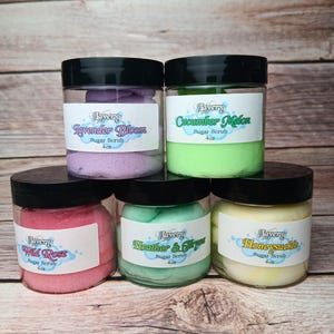 May include: Five glass jars with black lids filled with colorful sugar scrubs. The labels on the jars read "Lavery" and list the scents: Lavender Bloom, Cucumber Melon, Wild Rose, Heather & Thyme, and Honeysuckle. Each jar is 4 oz.