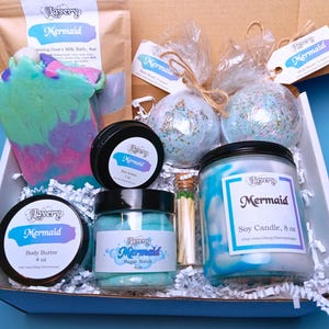May include: A gift box filled with bath and body products with a mermaid theme. The box contains a bath bomb, a sugar scrub, a body butter, a candle, and a bar of soap. The products are all in shades of blue and green.