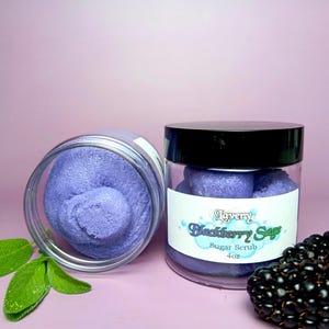 May include: Two jars of purple sugar scrub with a label that reads "Lavery Blackberry Sage Sugar Scrub 4 oz" and blackberries on a pink background.