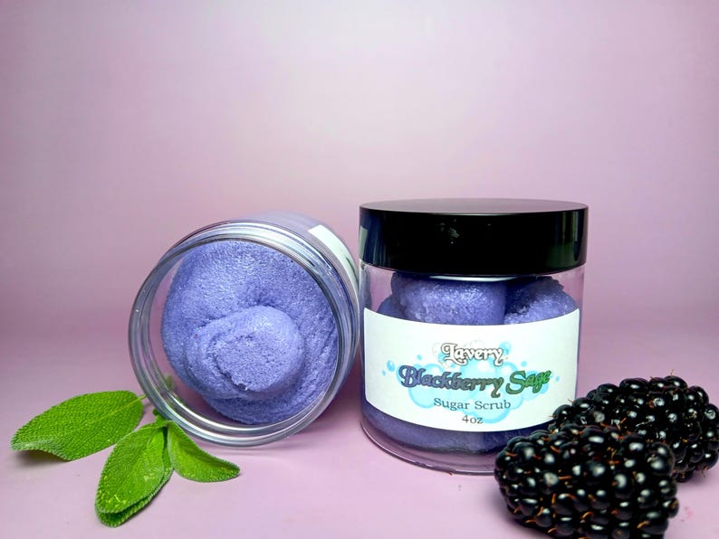 May include: Two jars of purple sugar scrub with a label that reads "Lavery Blackberry Sage Sugar Scrub 4 oz" and blackberries on a pink background.