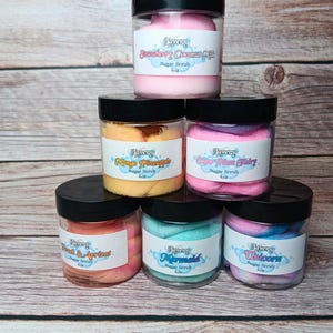 May include: Six glass jars of sugar scrub with colorful swirls of pink, blue, and yellow. The jars are labeled with the names "Strawberry Coconut Milk", "Mango Pineapple", "Sugar Plum Fairy", "Mermaid", "Unicorn", and "Peach & Apricot".