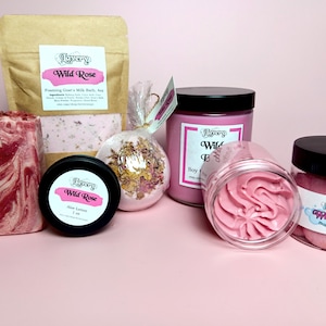 May include: A Wild Rose themed bath and body gift set with a pink and white marbled soap bar, a bath bomb, a jar of sugar scrub, a jar of whipped body butter, a jar of aloe lotion, and a soy candle.