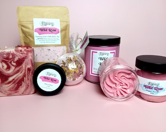 May include: A Wild Rose themed bath and body gift set with a pink and white marbled soap bar, a bath bomb, a jar of sugar scrub, a jar of whipped body butter, a jar of aloe lotion, and a soy candle.