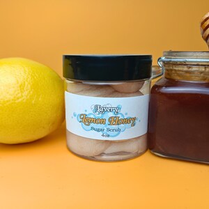 May include: A clear glass jar with a black lid filled with a light brown sugar scrub. The label reads "Lavery Lemon Honey Sugar Scrub 4 oz". A lemon and a jar of honey are also in the image.