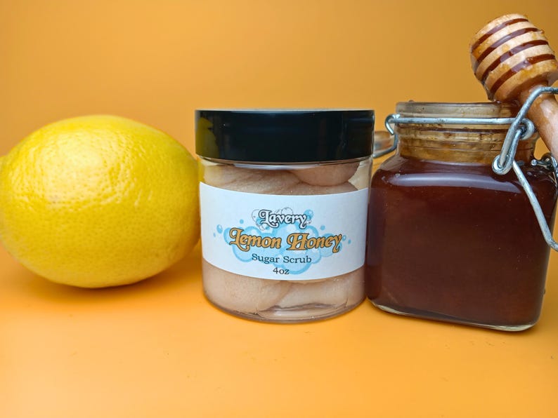 May include: A clear glass jar with a black lid filled with a light brown sugar scrub. The label reads "Lavery Lemon Honey Sugar Scrub 4 oz". A lemon and a jar of honey are also in the image.