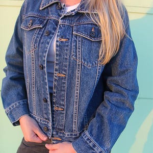 Puede incluir: Chaqueta vaquera azul clásica con botones, bolsillos en el pecho y cuello. La chaqueta tiene un aspecto vintage con un lavado medio, ideal para un look casual.