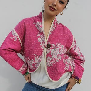 以下が含まれることがあります： 鮮やかなピンク色のキルティングジャケットで、白い花柄、ダークブラウンのボタン1つ留め、縁にはクリーム色のトリミングが施されています。ジャケットは長袖で、生地全体に白いステッチが見え、裾は丸みを帯びたデザインになっています。