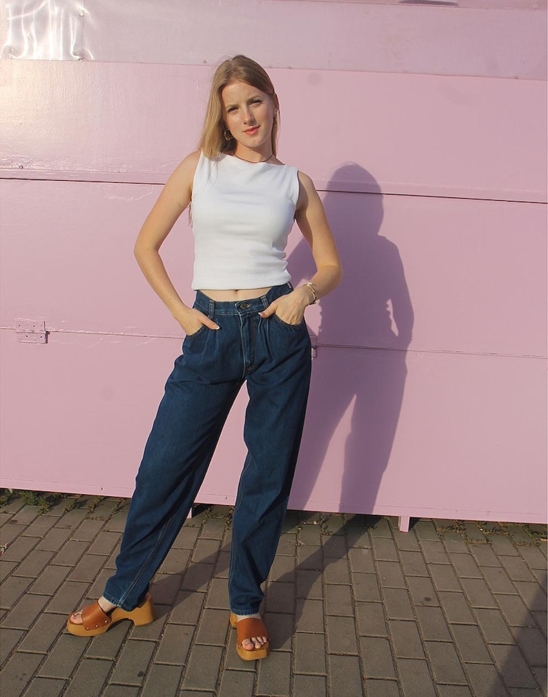 May include: A person wearing a white vest top and dark blue jeans with a high waist. They are wearing brown sandals with a wooden sole. The background is a pink wall.