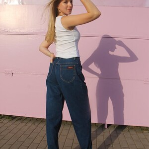 May include: A woman in a white vest top and dark blue jeans. The jeans are high-waisted with a small logo on the back pocket. She is also wearing brown wooden clogs. The background is a pink wall.