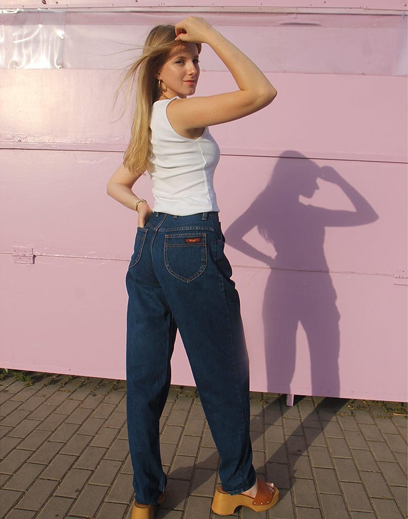 May include: A woman in a white vest top and dark blue jeans. The jeans are high-waisted with a small logo on the back pocket. She is also wearing brown wooden clogs. The background is a pink wall.