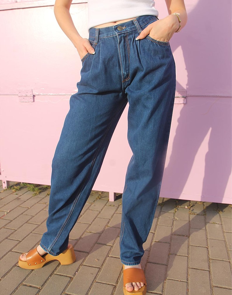 May include: Dark wash denim jeans with a high waist and pleated front. The jeans have a relaxed fit and are paired with brown platform sandals. The model is wearing a white top.