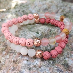 May include: Two beaded bracelets. One bracelet features pink lava rock beads and rose gold accents. The other bracelet has white and pink beads. The bracelets are on a stone surface.