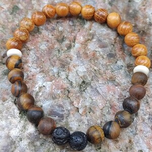 May include: A bracelet made of various beads, including wood, tiger eye, lava, and bone. The bracelet's colour scheme is natural, with shades of brown, tan, and black. The beads are arranged in a circular pattern.