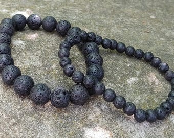 Op de afbeelding: Twee zwarte lavasteen kralenarmbanden. De armbanden zijn gemaakt van ronde, poreuze kralen. De ene armband is groter dan de andere, met grotere kralen. De armbanden liggen op een grijs, getextureerd oppervlak.