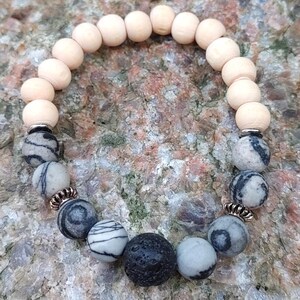 May include: A bracelet made of mixed beads, including light-coloured wooden beads, patterned grey and white stone beads, and a black lava stone bead. The bracelet is accented with copper-coloured metal spacers. The beads are strung together.