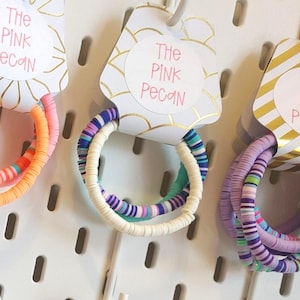 Peut inclure: Bracelets de perles colorées dans diverses nuances de rose, orange, vert, violet et bleu sont exposés sur un panneau perforé blanc. Chaque bracelet est attaché à une étiquette indiquant "The Pink Pecan".