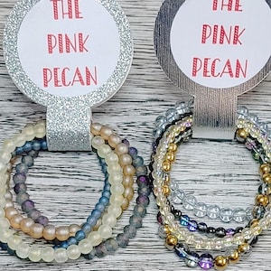 May include: Two stacks of beaded bracelets are displayed on decorative holders. The holders have the text "THE PINK PECAN" in red. One stack features blue, purple, and cream-coloured beads. The other stack has gold, clear, and iridescent beads.