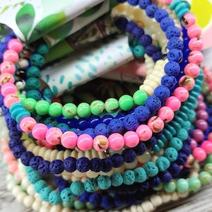 May include: A collection of vibrant beaded bracelets in shades of pink, blue, green, and cream. The bracelets feature lava beads and round beads. The packaging displays the text "The Pink Pecan".
