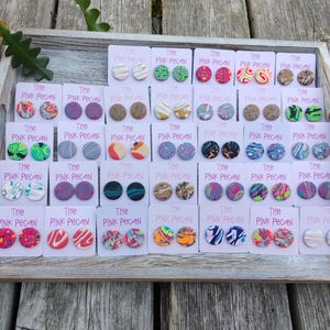 May include: A collection of colourful stud earrings presented on white cards within a wooden tray. The earrings showcase diverse patterns and hues, such as swirls, dots, and marbled effects. The cards are labelled "The Pink Pecan."