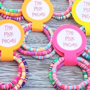 May include: A wooden tray displays multiple colourful bracelets, each with a tag bearing the text "The Pink Pecan". The bracelets are composed of small, brightly coloured beads in shades of pink, yellow, blue, and green.