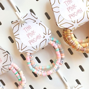 May include: A collection of colourful beaded bracelets, each with a tag reading "The Pink Pecan." The bracelets display pastel shades of pink, blue, and yellow. They are presented on a white pegboard.