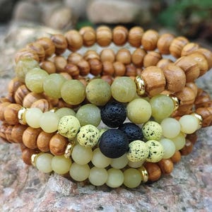 May include: A collection of beaded bracelets in shades of brown, green, and cream. The bracelets feature wooden beads, green stone beads, black lava beads, and cream-coloured beads with gold accents. The bracelets are stacked together.