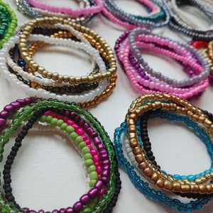 May include: A collection of colourful beaded bracelets in various shades of green, pink, purple, blue, gold, and black. The bracelets are stacked on a white surface.