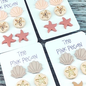 May include: Four sets of seashell, starfish, and sand dollar shaped earrings on white cards. The earrings are in shades of pink, tan, and beige. The text "The PINK Pecan" is written on each card in purple ink.
