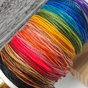 May include: A spool of thin, twisted cord featuring a vibrant gradient of colours including black, brown, red, pink, orange, yellow, green, blue, and purple. The cord is tightly wound in parallel rows, creating a continuous spectrum of hues.