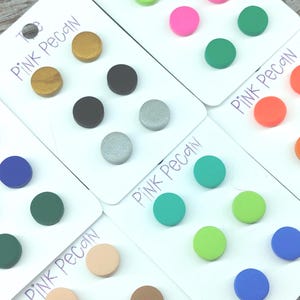 May include: A selection of colourful stud earrings displayed on white cards. The earrings come in a variety of colours, including gold, black, silver, pink, green, and blue. The cards are labelled "Pink Pecan" in handwritten script.