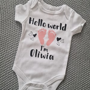 Può includere: Body da bambino bianco con la scritta nera "Hello world I'm Oliwia". Il body presenta impronte di piedi rosa e grafiche a cuore nere. L'indumento è realizzato in materiale morbido e ha chiusure a scatto.