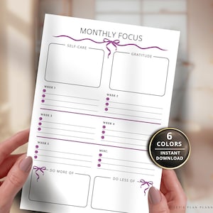 May include: A white planner page with the text "MONTHLY FOCUS" and sections for self-care, gratitude, and weekly goals. The planner includes the text "6 COLORS" and "INSTANT DOWNLOAD". The planner is held by a person.