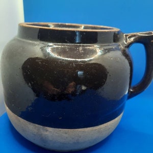May include: A black and white glazed ceramic crock with a handle. The crock has a rounded body and a wide mouth.