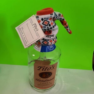 May include: A clear glass bottle with a white and orange knitted bottle topper. The bottle is labeled "Tito's Handmade Vodka" and has a blue label with "Texas USA Bottled" printed on it.