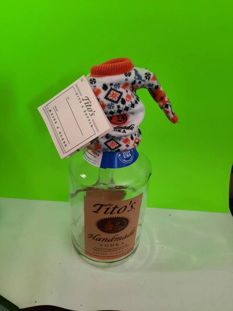 May include: A clear glass bottle with a white and orange knitted bottle topper. The bottle is labeled "Tito's Handmade Vodka" and has a blue label with "Texas USA Bottled" printed on it.