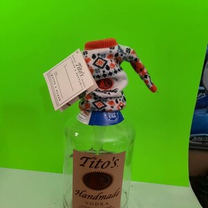 May include: A clear glass bottle of Tito's Handmade Vodka with a white and orange knitted bottle cozy. The bottle has a brown label with the text "Tito's Handmade Vodka" and "Austin, Texas".