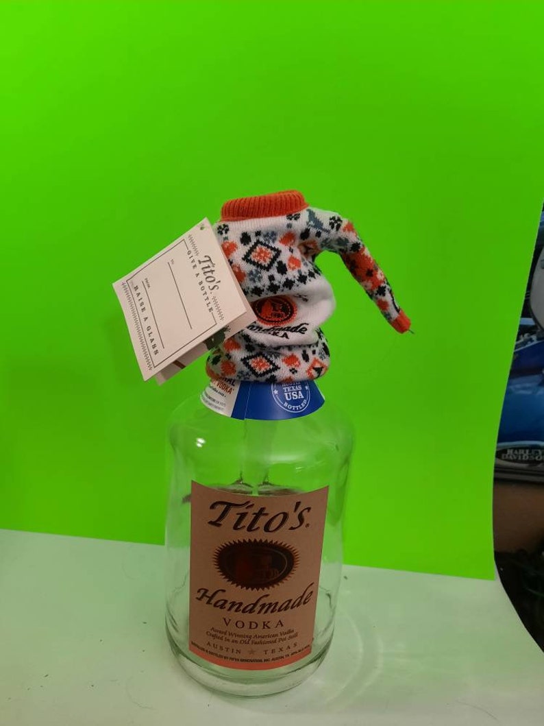 May include: A clear glass bottle of Tito's Handmade Vodka with a white and orange knitted bottle cozy. The bottle has a brown label with the text "Tito's Handmade Vodka" and "Austin, Texas".